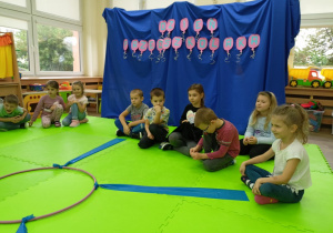 Dzieci siedzące na dywanie, w tle dekoracja z napisem "Dzień Przedszkolaka".